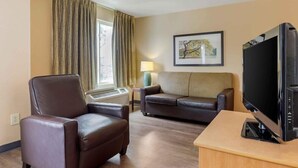 Habitación de hotel con un sillón de cuero marrón, un sofá, un televisor sobre un mueble, una lámpara y un cuadro en la pared.