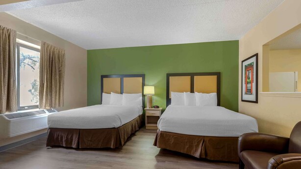 Una habitación de hotel con dos camas, una ventana con cortinas, un calefactor, una mesita de noche con lámpara y un sillón de cuero marrón.