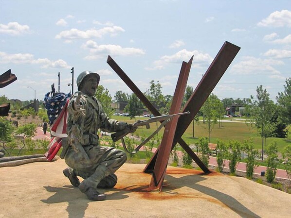 A soldier in camouflage kneeling with a rifle, holding an American flag, in an outdoor setting with metal structures and a clear sky. - Ramada Hotel & Conference Center by Wyndham Hammond (Hammond)