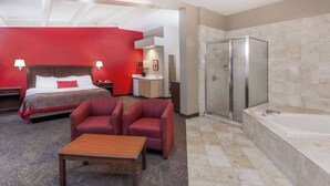 A hotel room with a bed, two armchairs, a wooden table, a bathroom with a bathtub, and a shower. - Ramada Hotel & Conference Center by Wyndham Hammond (Hammond)