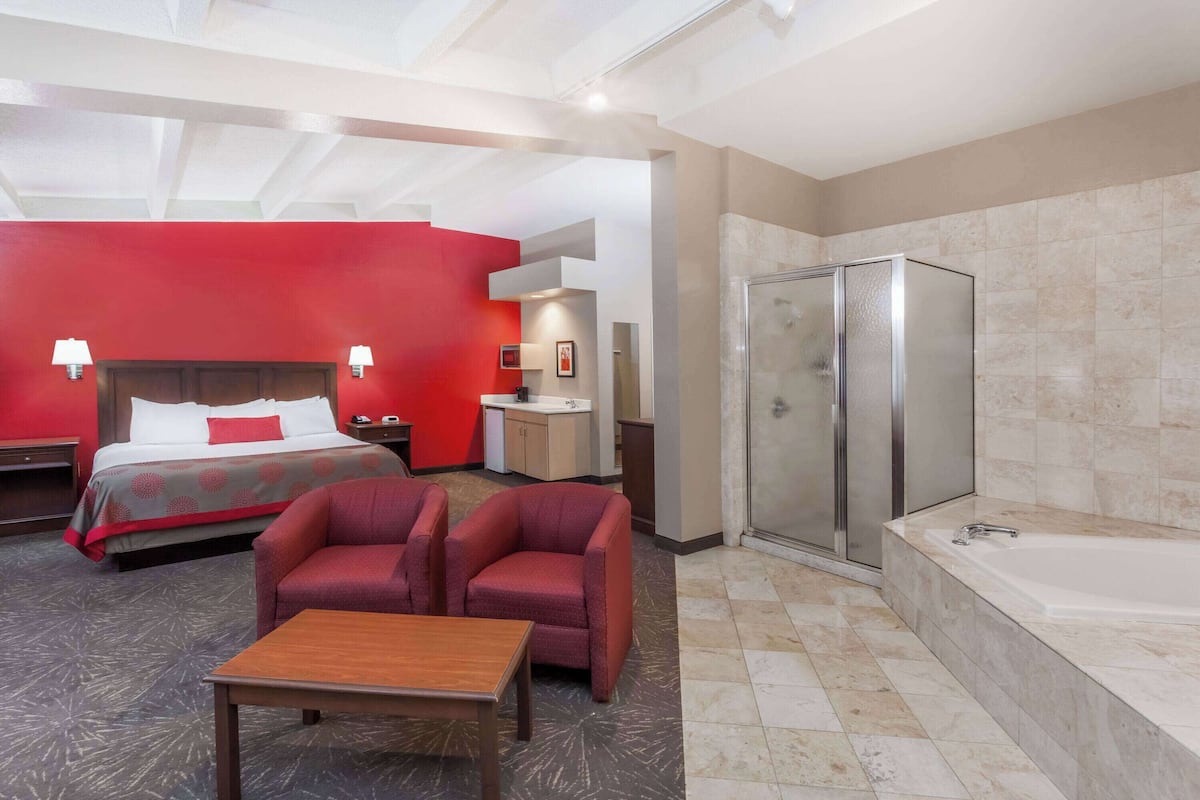 A hotel room with a bed, two armchairs, a wooden table, a bathroom with a bathtub, and a shower.