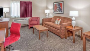 A hotel room with a red chair, a brown sofa, two side tables with lamps, a flat-screen TV, and a large window with curtains. - Ramada Hotel & Conference Center by Wyndham Hammond (Hammond)