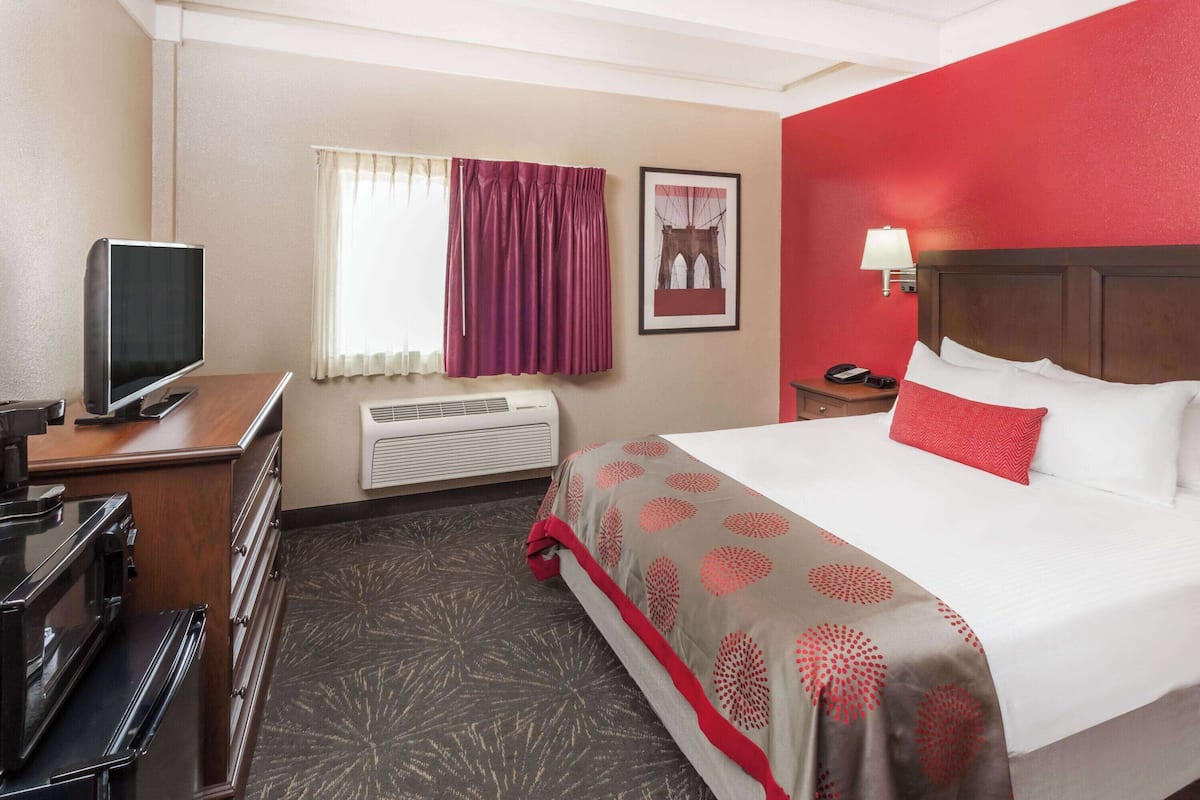 A hotel room with a bed, a desk with a television, an air conditioner, a window with curtains, and a wall-mounted lamp.