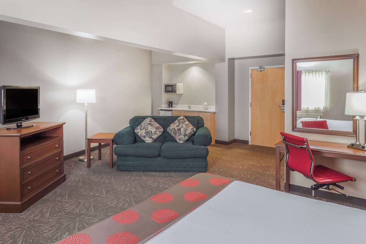 A hotel room with a TV, sofa, desk, chair, and a small kitchenette.