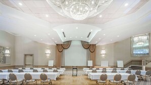 A conference room with a high ceiling, chandeliers, and a stage with curtains. Tables and chairs are arranged for meetings. - Ramada Hotel & Conference Center by Wyndham Hammond (Hammond)