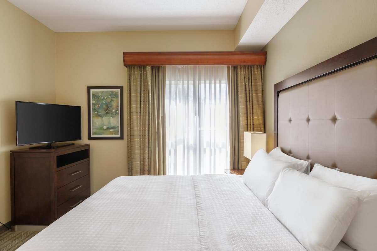Ein Hotelzimmer mit einem großen Bett, einem Flachbildfernseher, einer Kommode und einem Fenster mit Vorhängen.