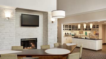 A modern hotel room with a fireplace, a flat-screen TV mounted on the wall, a dining area with a large table and chairs, and a kitchen area with a bar and counter.
