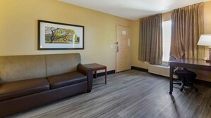 Une chambre d’hôtel avec un canapé en cuir marron, une petite table en bois, un bureau avec une chaise, un tableau encadré au mur et une fenêtre avec des rideaux.