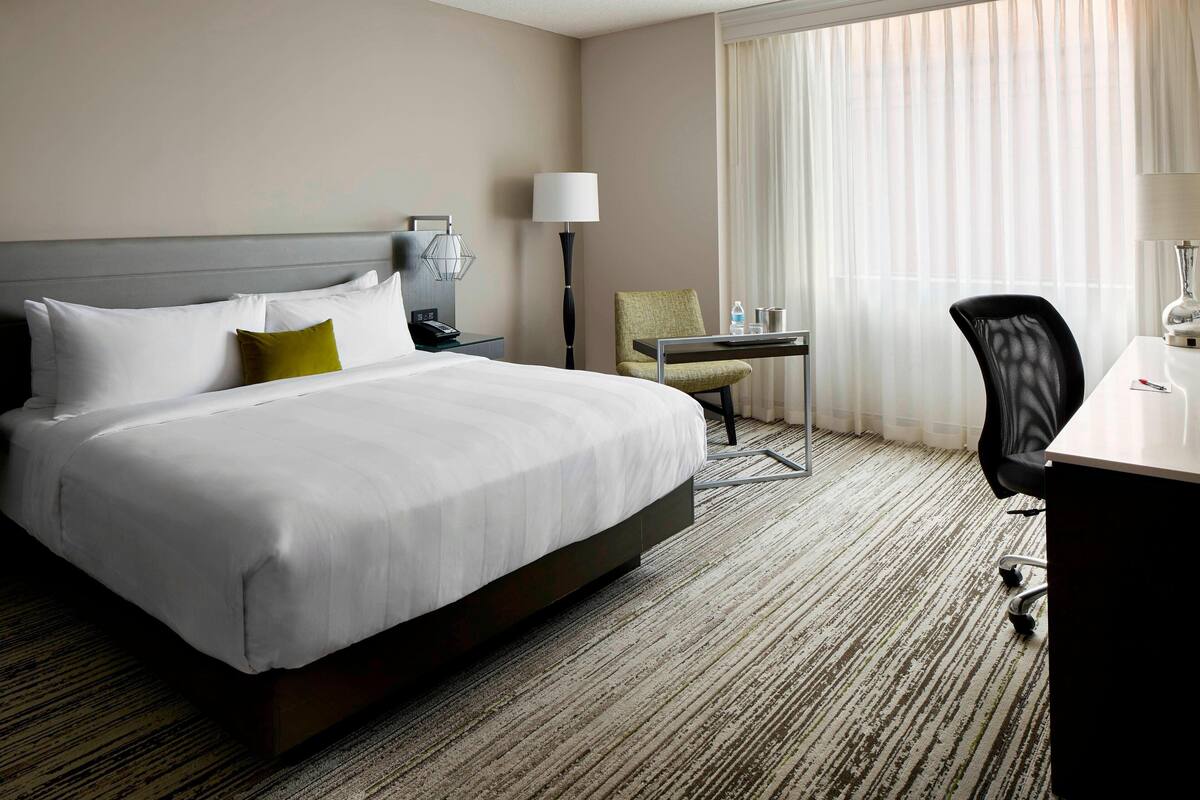 A hotel room with a large bed, a desk, a chair, a lamp, and a window with curtains.