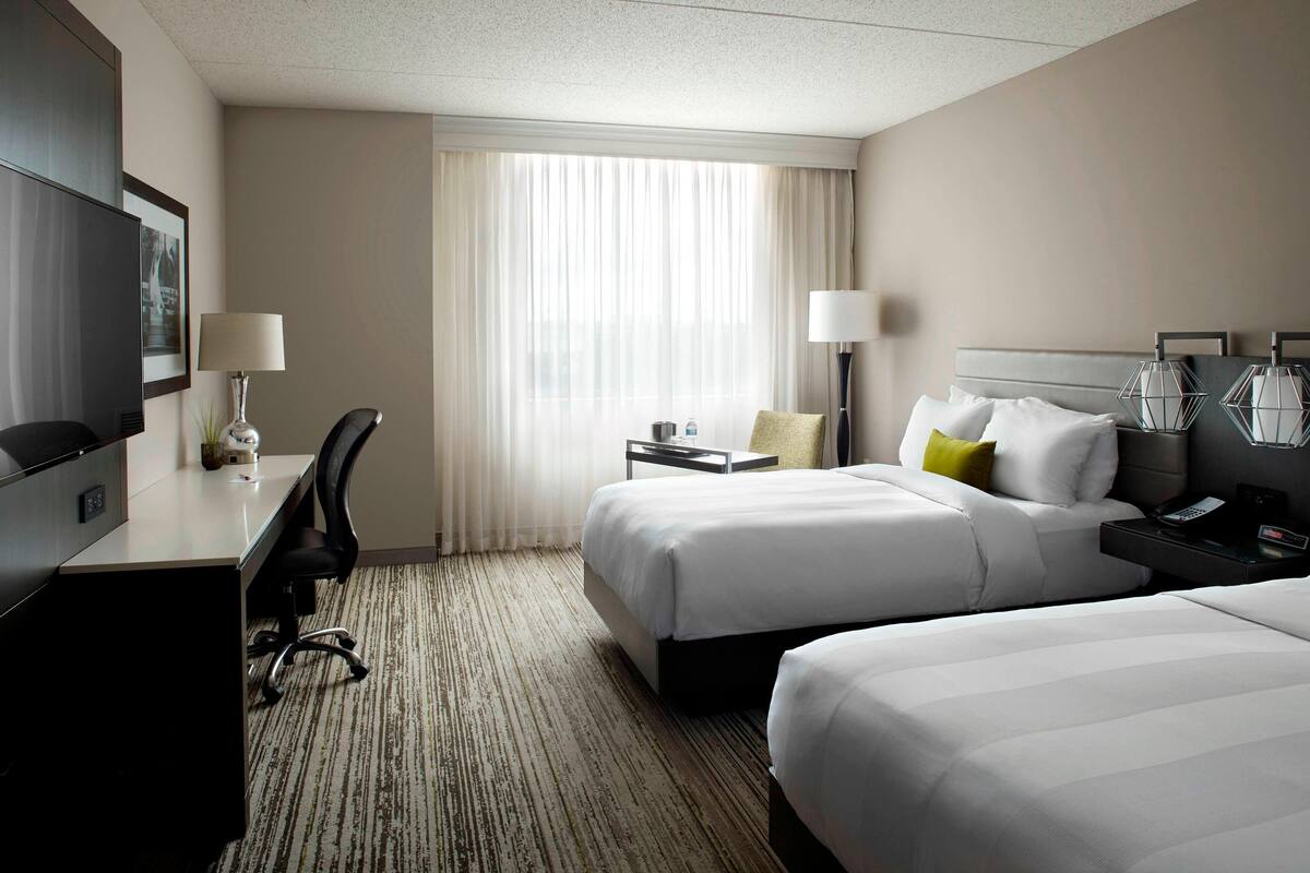 A hotel room with two beds, a desk, a chair, a TV, and a window with curtains.