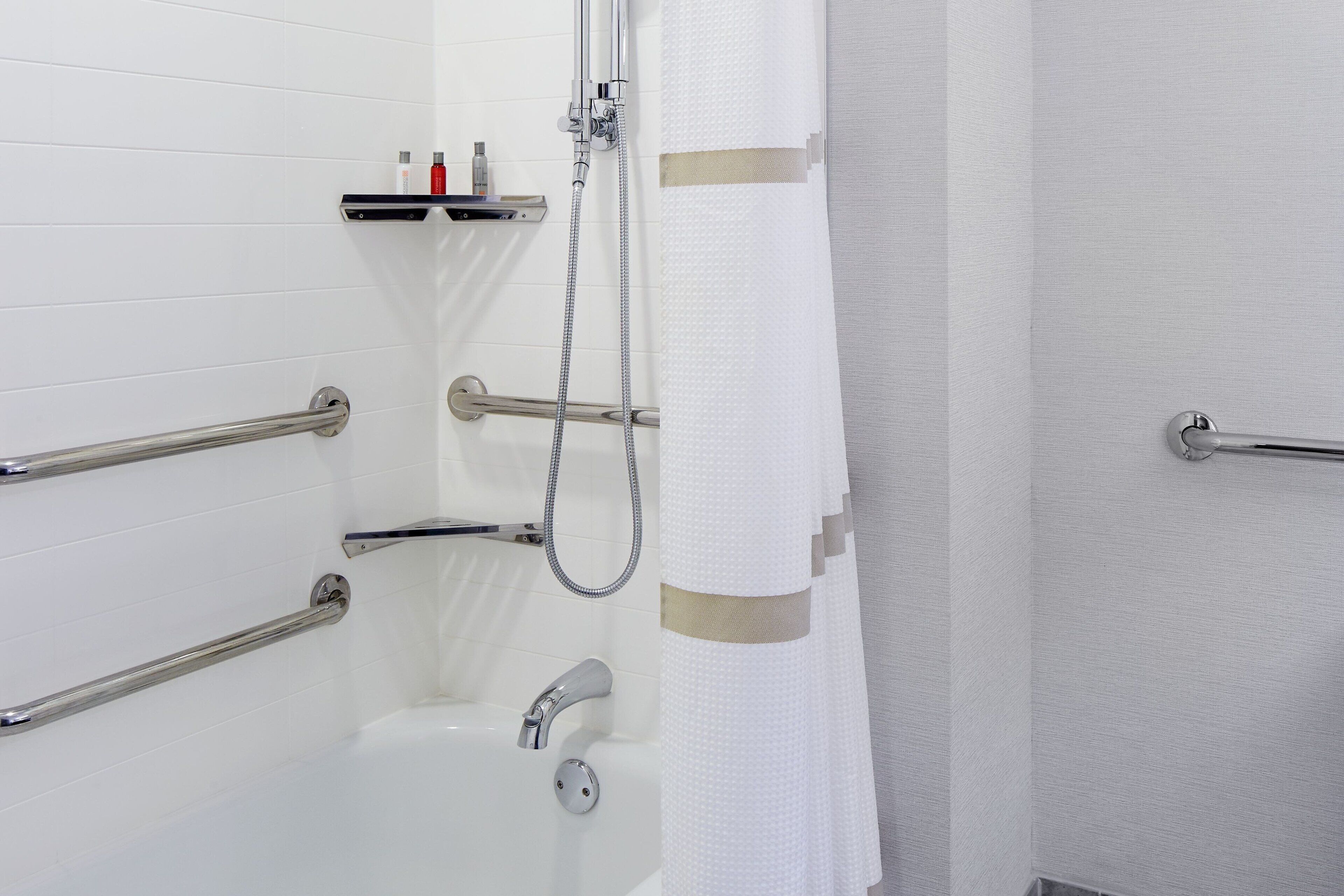 A modern bathroom with white tiled walls, a bathtub, and a shower with a handheld hose. There are grab bars installed for safety.