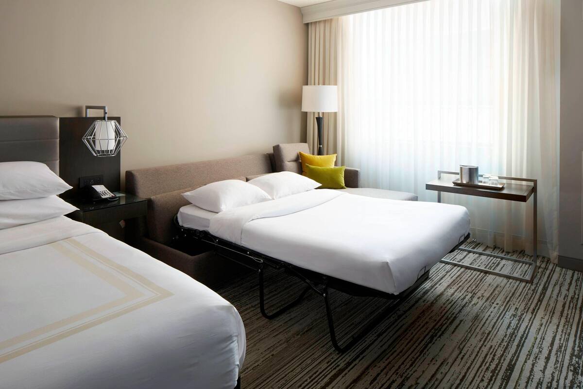 A hotel room with two beds, a bedside table with a lamp, a small table with a cup, and a window with curtains.