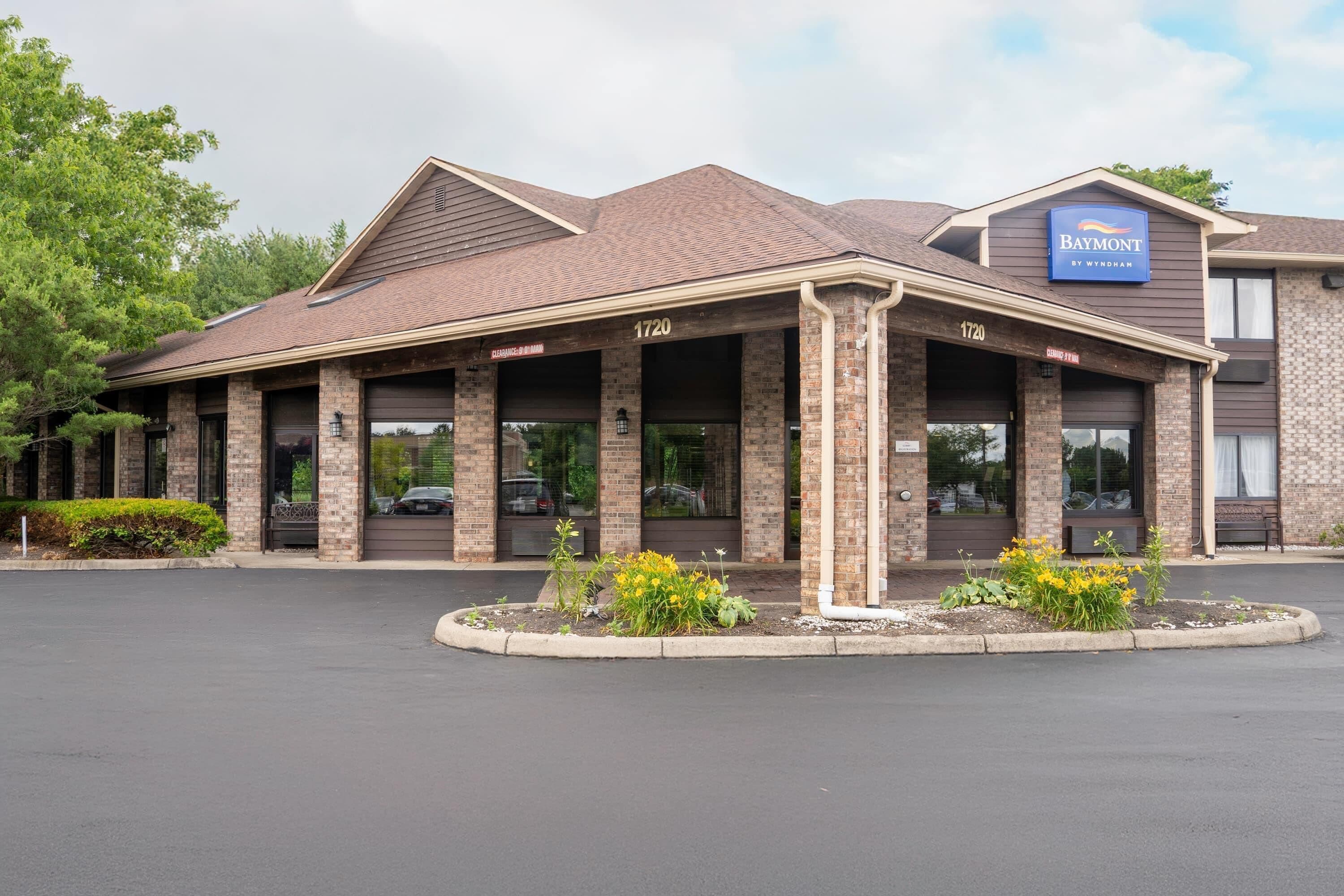 A Baymont by Wyndham hotel with a brick facade, numbered 1720, featuring a covered entrance and a landscaped area with yellow flowers.