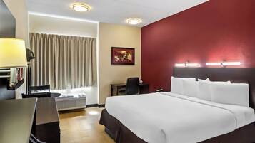 A hotel room with a large bed, a desk, a chair, a lamp, a picture on the wall, and a window with curtains.