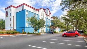 A multi-story hotel with a blue and white facade, a red roof, and a parking lot with cars.