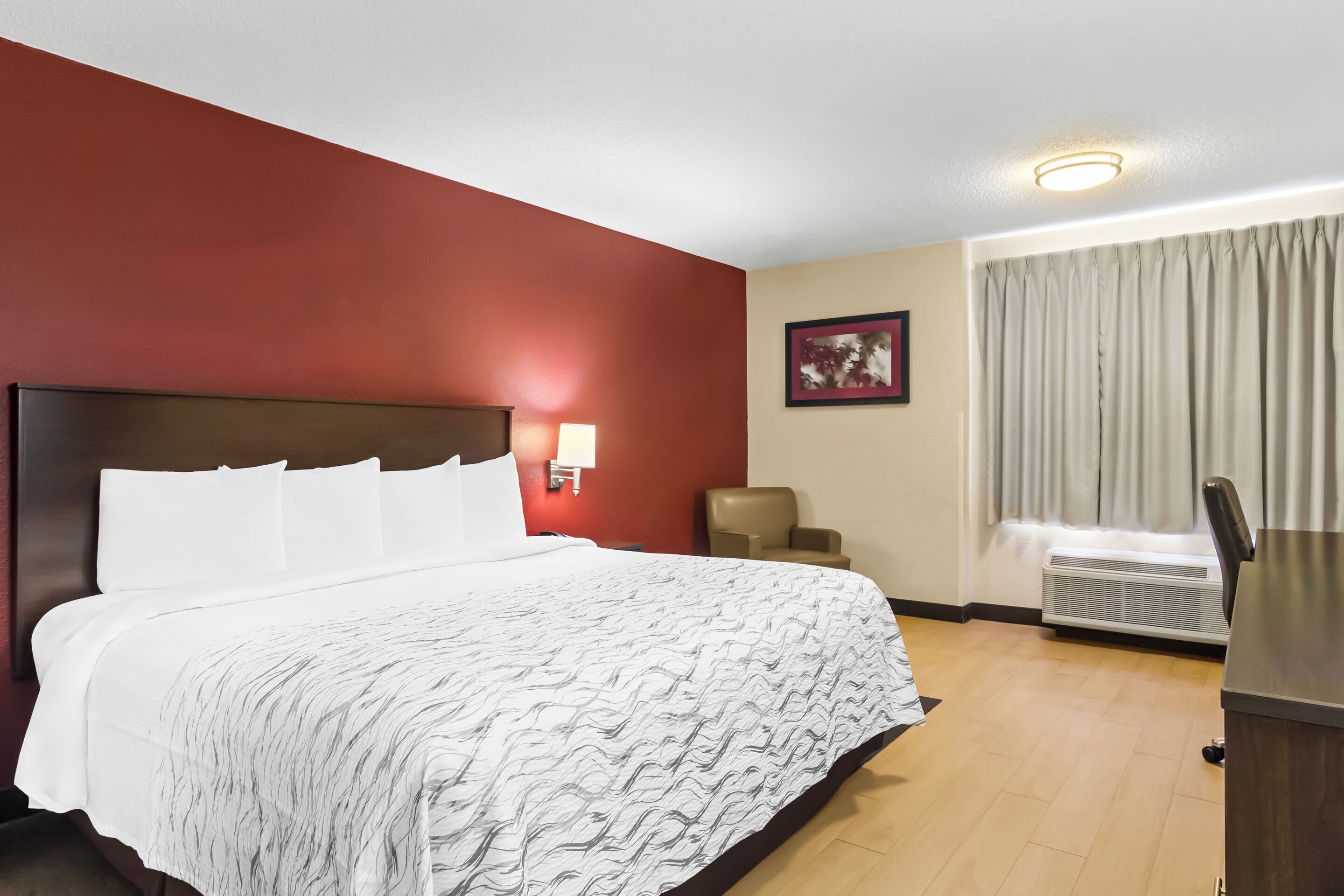 Habitación de hotel con una cama grande, un escritorio, una silla, una lámpara de pared y un ventanal con cortinas.