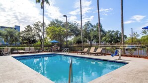A rectangular pool with a metal safety railing, surrounded by lounge chairs and umbrellas, with trees and a fence in the background.