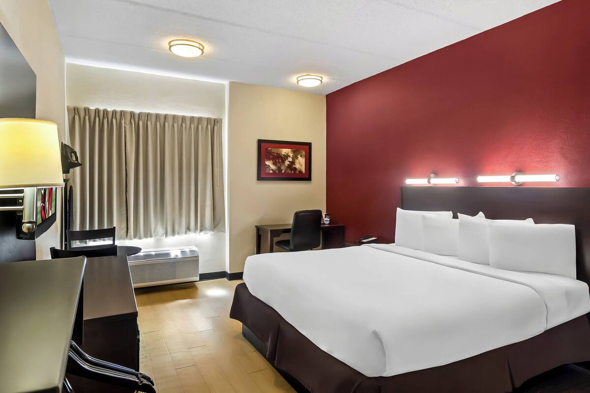 Ein Hotelzimmer mit einem großen Bett, einem Schreibtisch, einem Sessel, einer Lampe, einem Bild an der Wand und einem Fenster mit Vorhängen.
