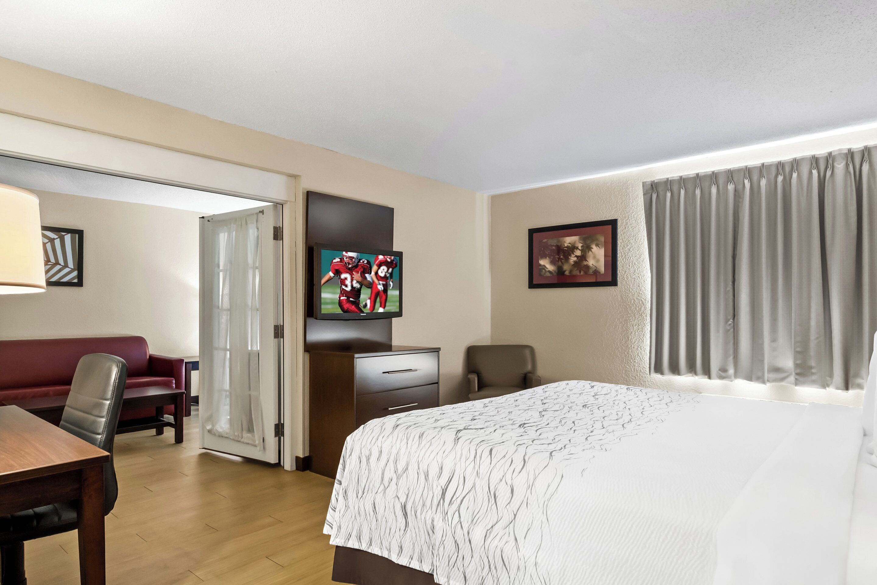 A hotel room with a bed, a desk, a chair, a TV, and a door leading to another room.