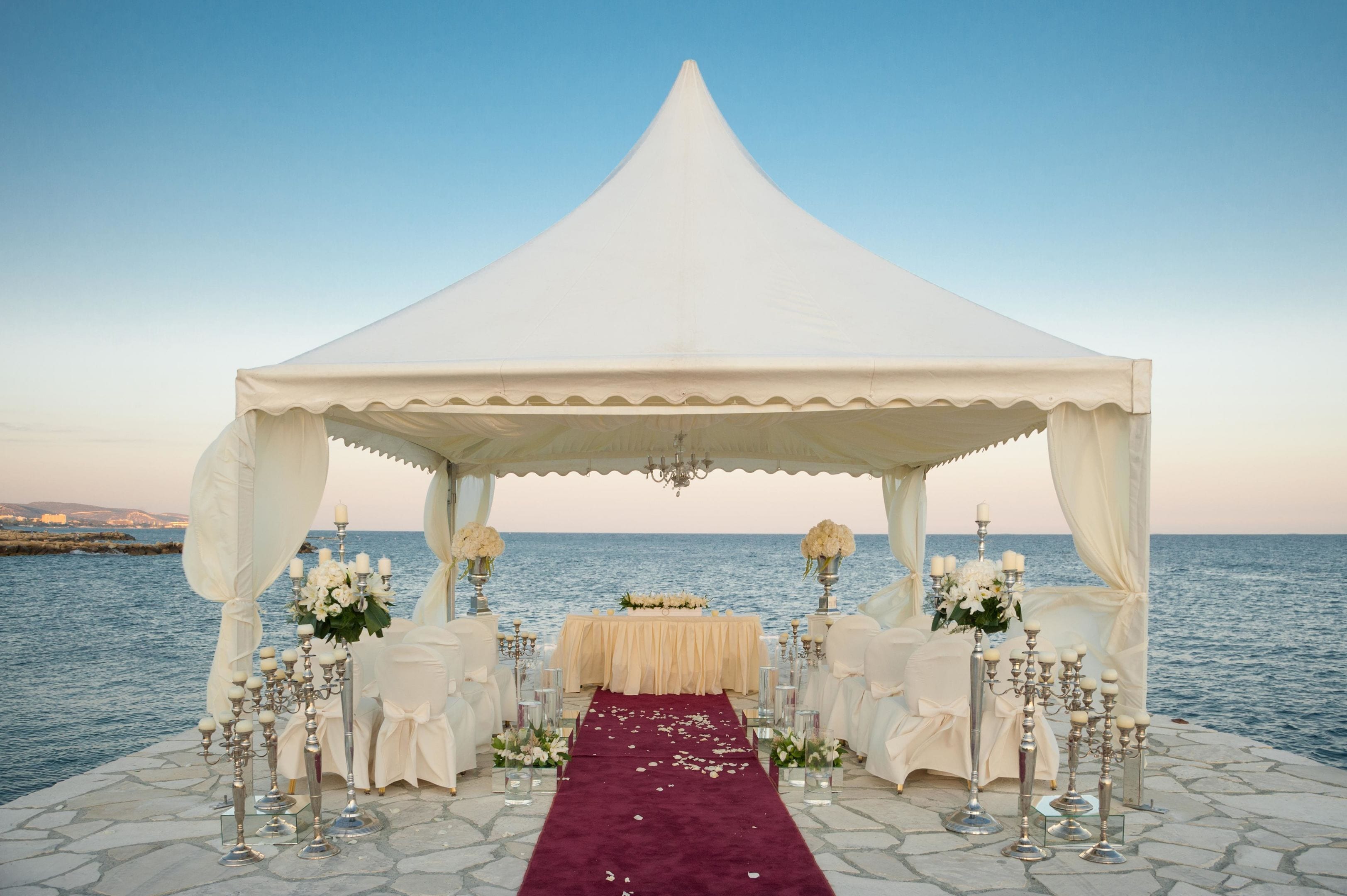 A white tented wedding setup by the sea with a red carpet, white chairs, and floral arrangements.