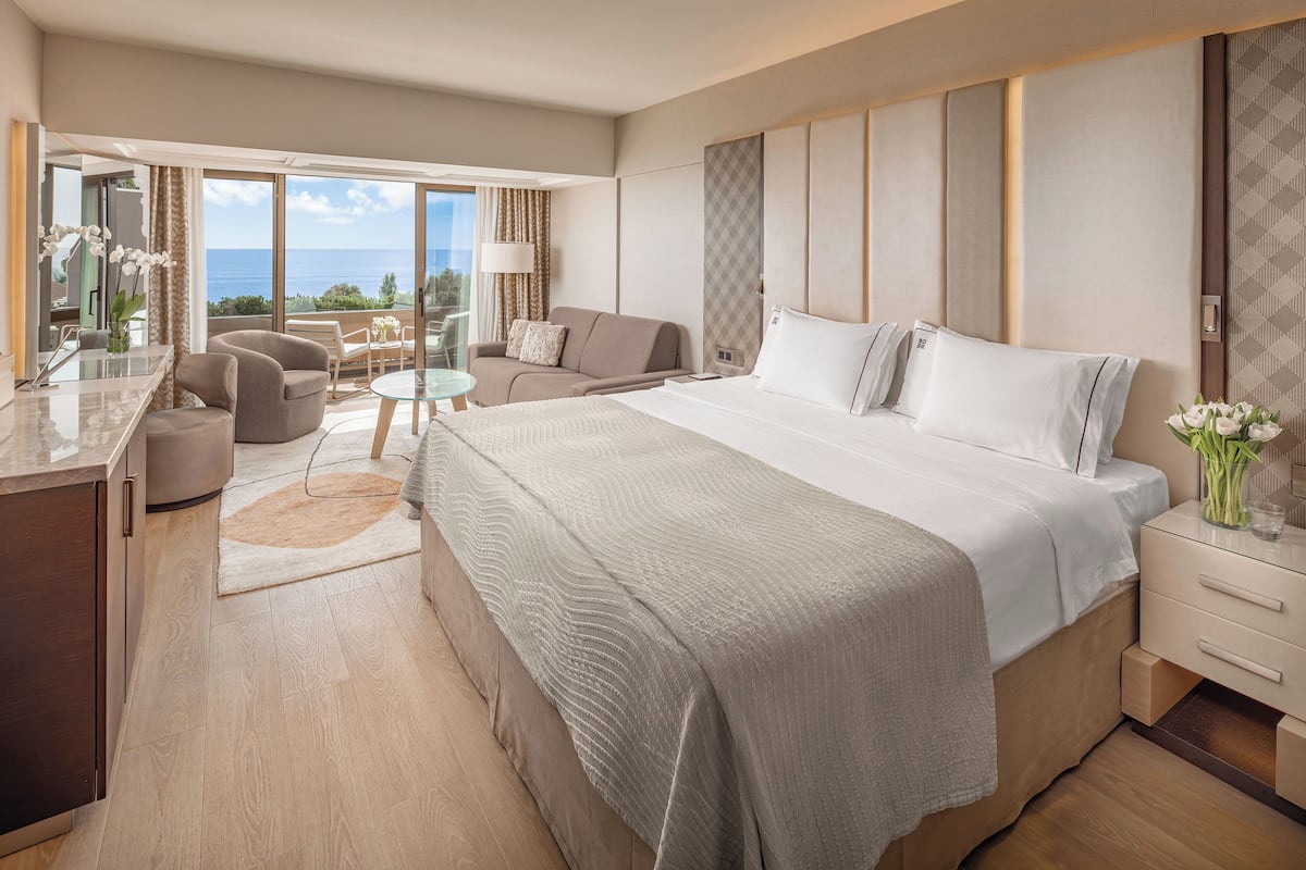 A modern hotel room with a large bed, a sitting area with a glass table, and a view of the ocean.