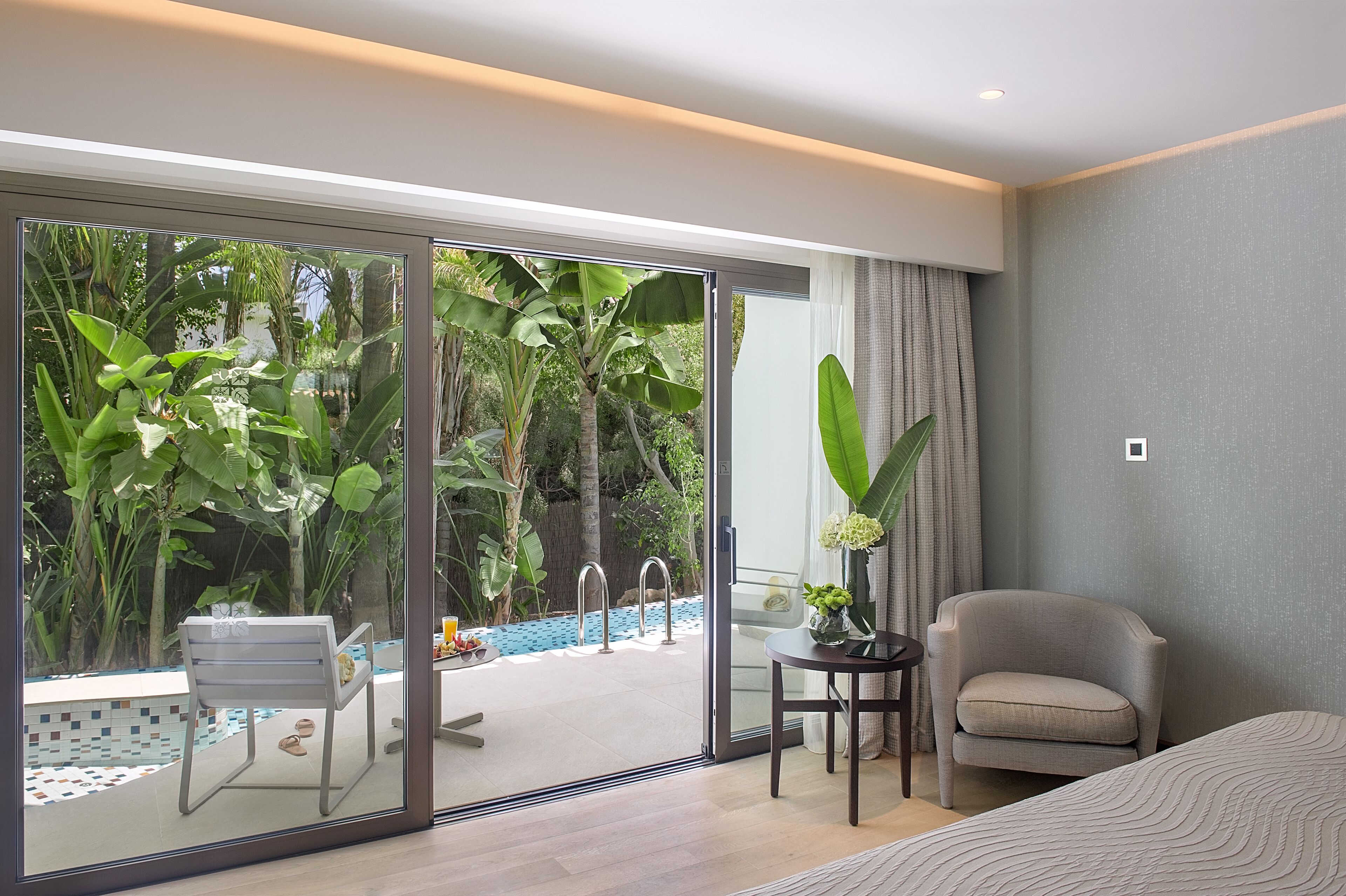 A modern hotel room with a sliding glass door leading to a pool area, a comfortable chair, and a small table with a plant.