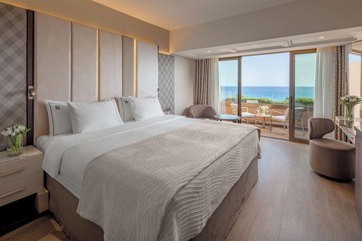 A hotel room with a large bed, a seating area with a view of the sea, and a balcony with a table and chairs.