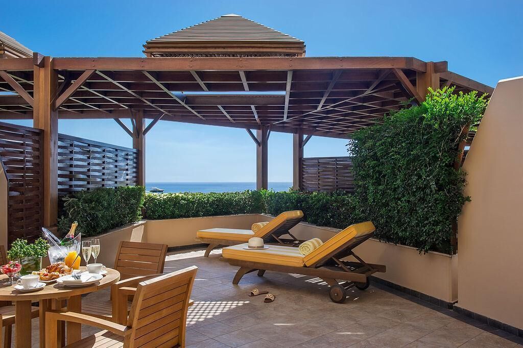 A covered outdoor seating area with lounge chairs and a table set for breakfast, overlooking the sea.