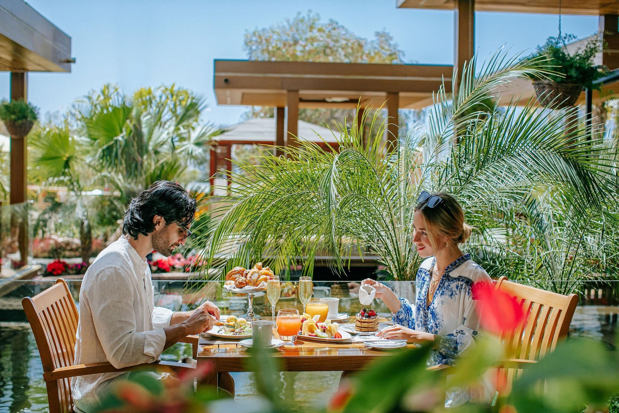A couple dining at an outdoor table with a view of a water feature and tropical plants.