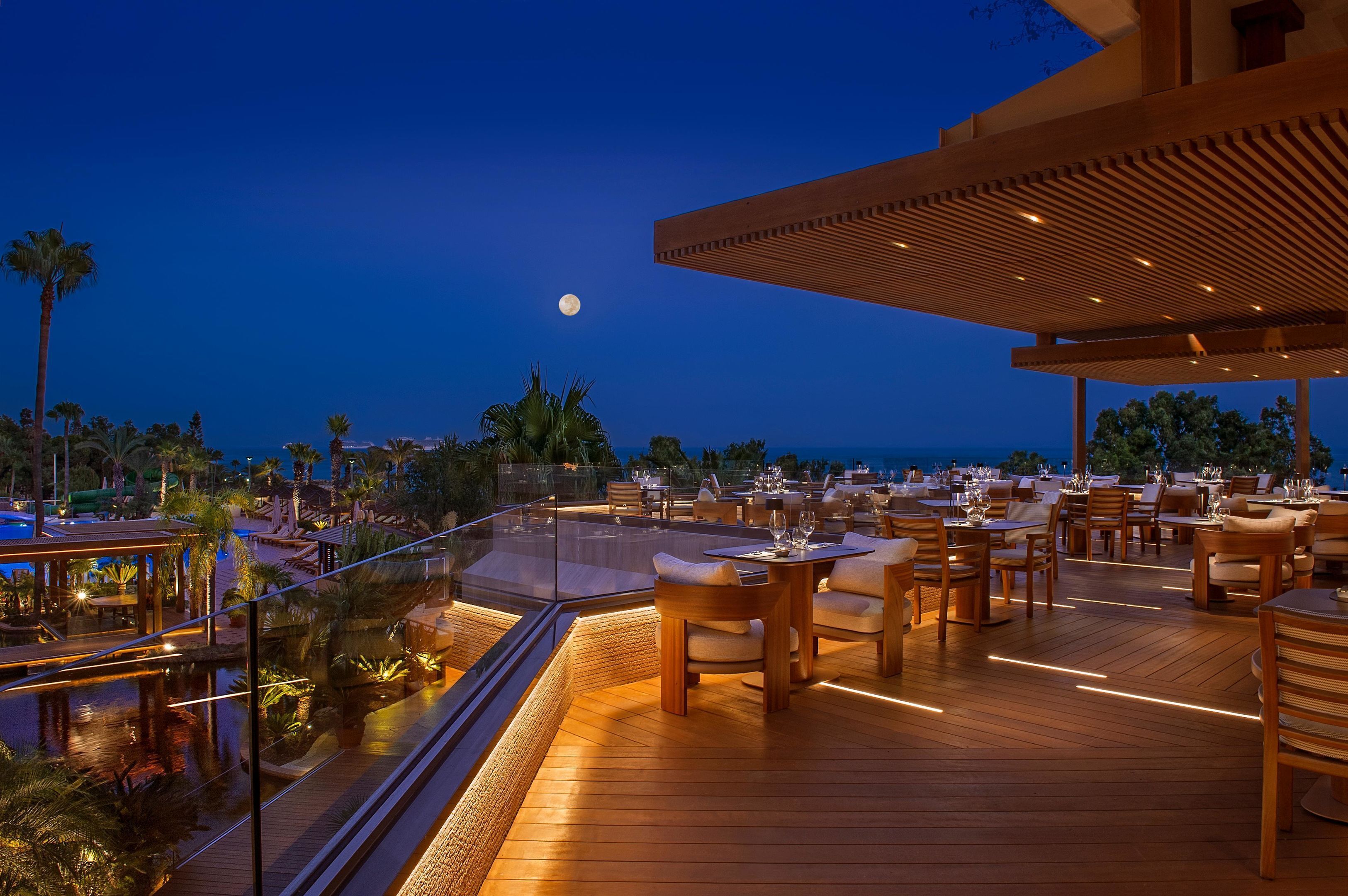 Zona de comedor en la azotea con mesas y sillas de madera, con vistas a una piscina y palmeras, bajo un cielo nocturno con luna visible.