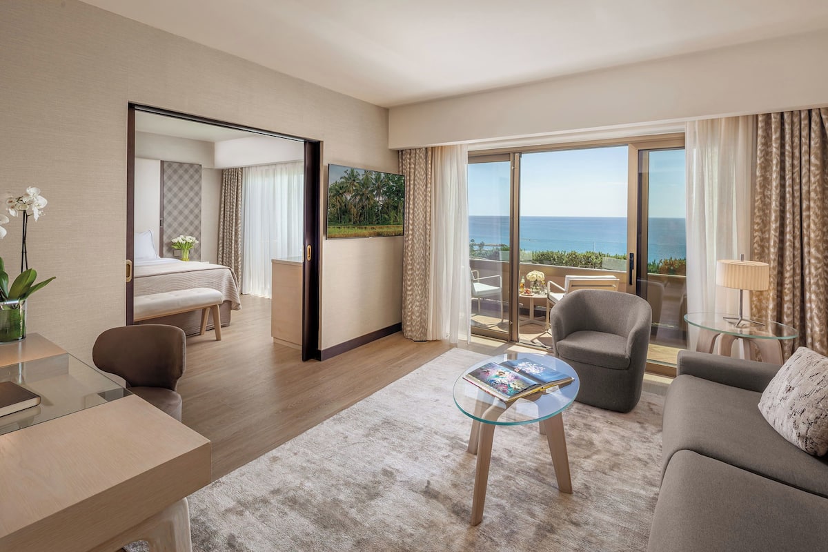 A modern hotel room with a balcony, a bed, a sitting area with a glass coffee table, and a desk.