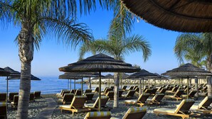 A beach with rows of wooden lounge chairs and thatched umbrellas, palm trees, and a clear blue sky. - Four Seasons Hotel (Limassol)