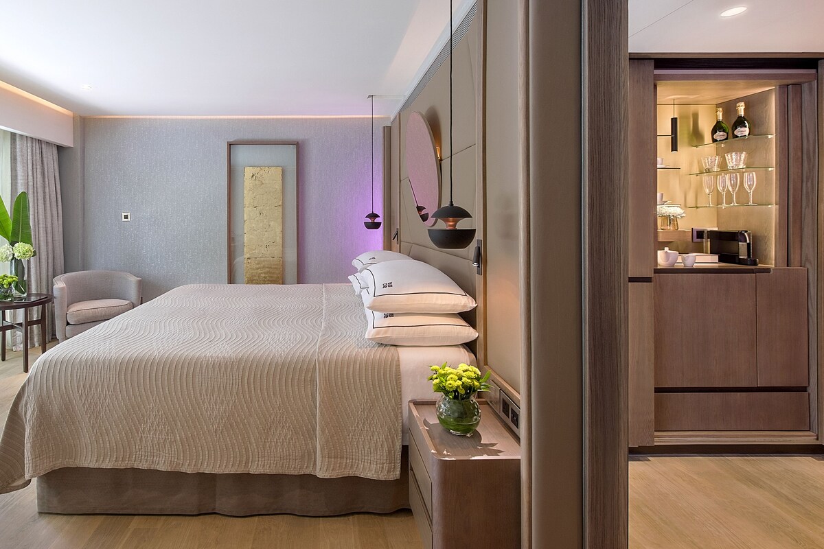 Une chambre d’hôtel moderne avec un grand lit, une table de chevet ornée d’un vase de fleurs, une chaise et une vue sur la salle de bain, qui comprend une coiffeuse et des étagères.