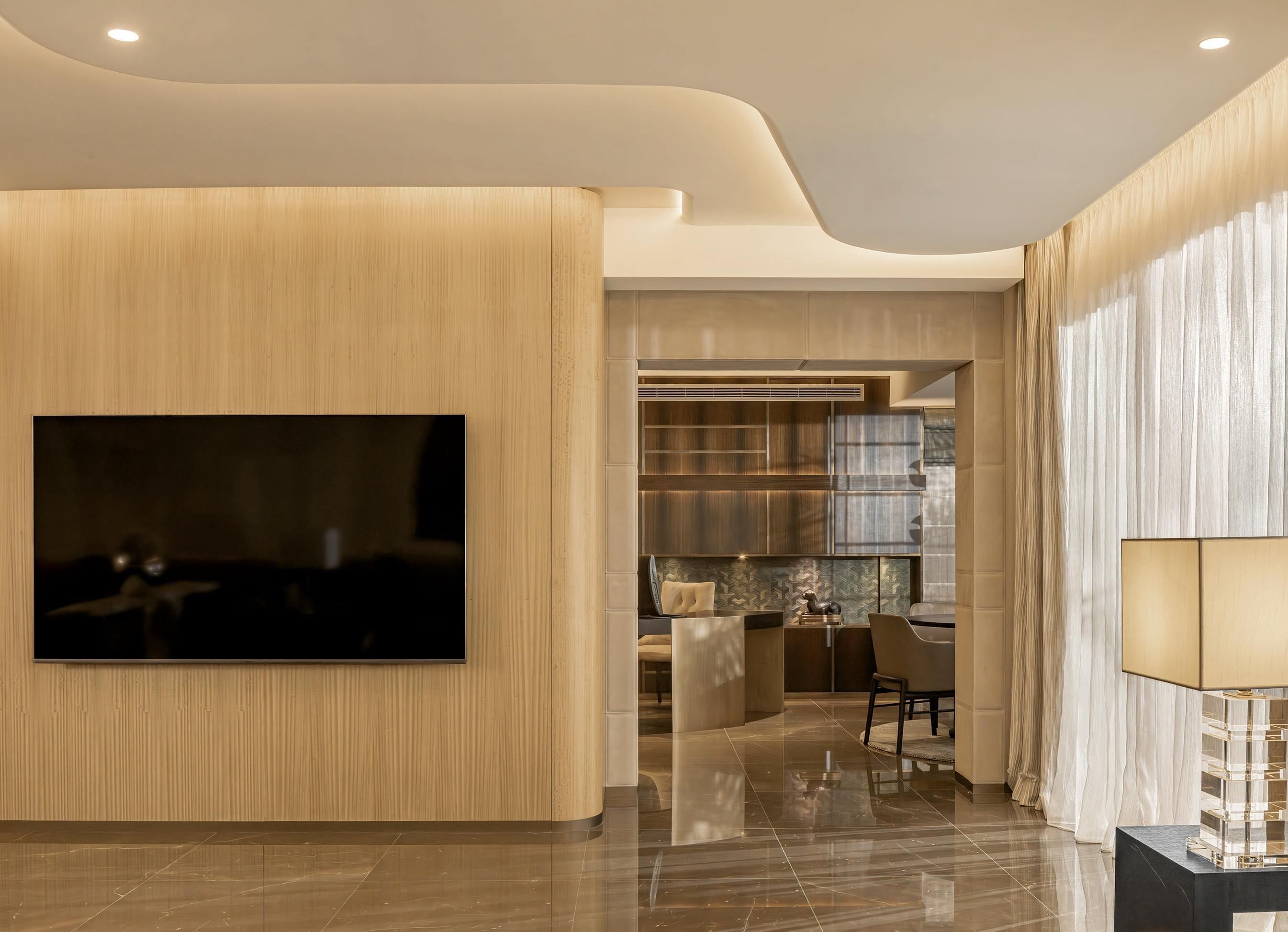 A modern interior with a large flat-screen TV mounted on a wooden wall, a sleek dining area with a chandelier, and a well-lit space with a lamp and a glass display.