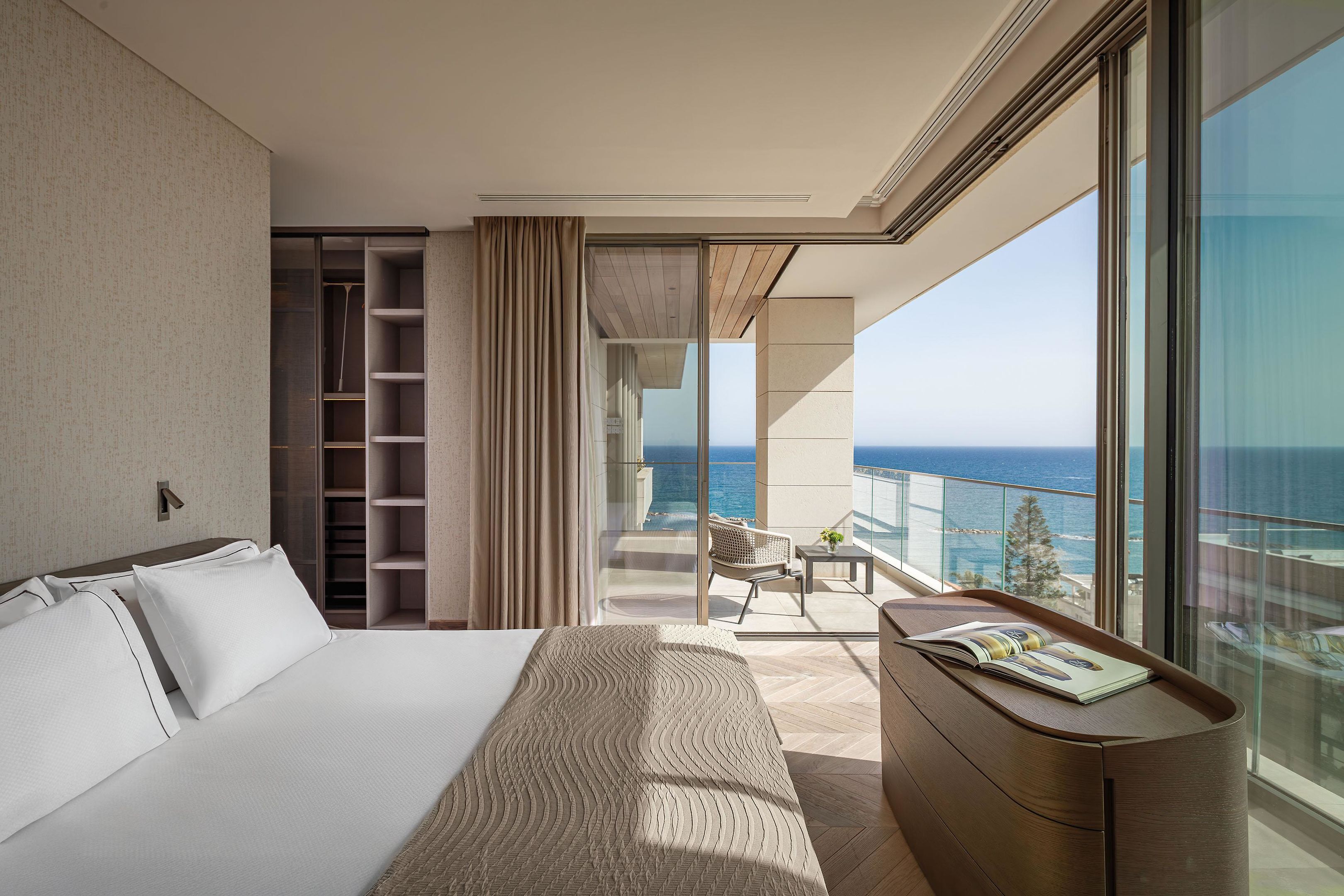 Una habitación de hotel moderna con una cama grande, un vestidor, un balcón con vistas al mar y un pequeño rincón de descanso.