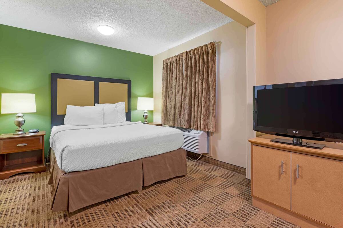 A hotel room with a bed, two bedside lamps, a television on a stand, and a window with curtains.