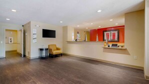 A spacious lobby with a wooden floor, a yellow armchair, a flat-screen TV mounted on the wall, and a red accent wall with a mirror and a small table with a laptop. - Extended Stay America Select Suites - Atlanta - Vinings (Atlanta)