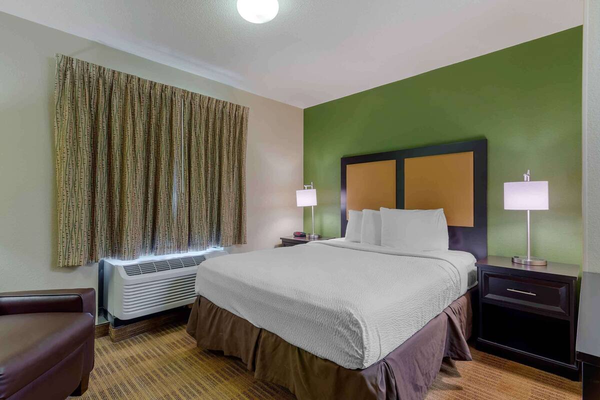 A hotel room with a large bed, two bedside tables with lamps, a chair, a large window with curtains, and an air conditioning unit.