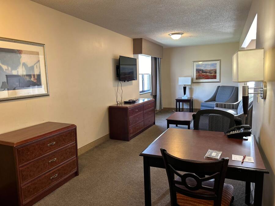 A hotel room with a wooden desk, a chair, a dresser, a television, a lamp, and two framed pictures on the walls.