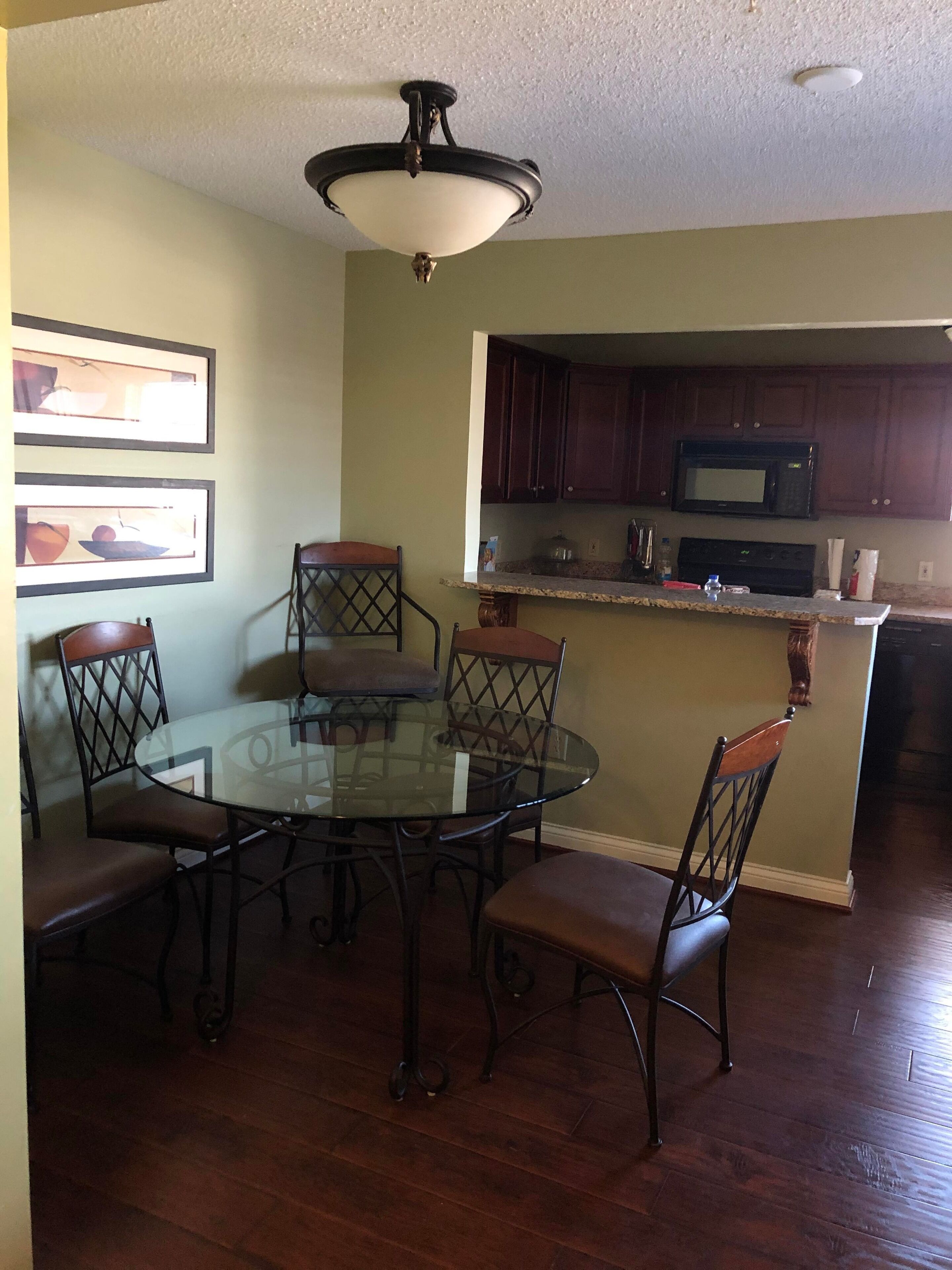 A dining area with a round glass table and six chairs, a kitchen with dark wooden cabinets and a microwave, and a ceiling light fixture.