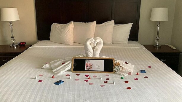 Una cama bien hecha con sábanas blancas, cabecera de madera y una decoración de toallas y confeti con forma de corazón sobre la cama.