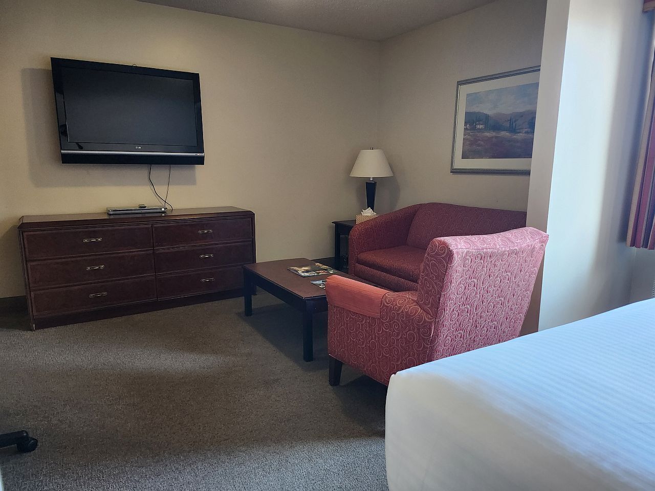 Habitación de hotel con cama, televisión montada en la pared, escritorio con lámpara, silla y cómoda.