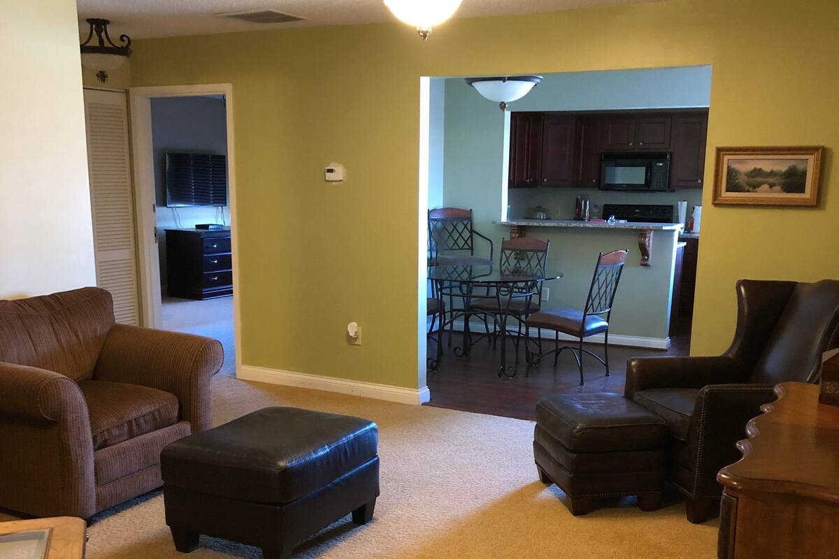 A living room with a brown sofa, ottoman, and wooden side table. There is a kitchen visible through an open doorway.