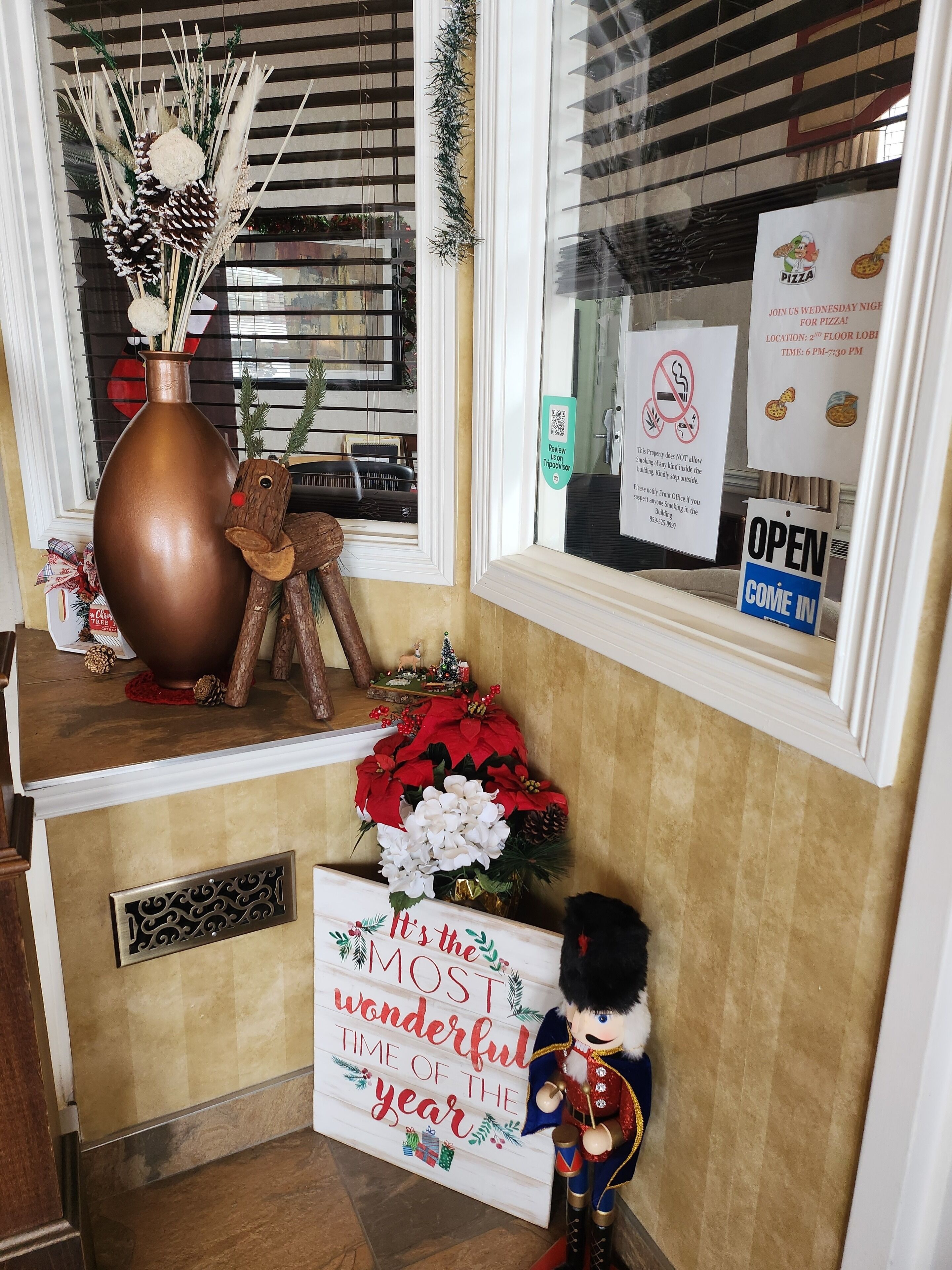 Uma entrada decorada com um vaso grande, um ursinho de pelúcia e uma placa com a mensagem 'É a época mais maravilhosa do ano'.