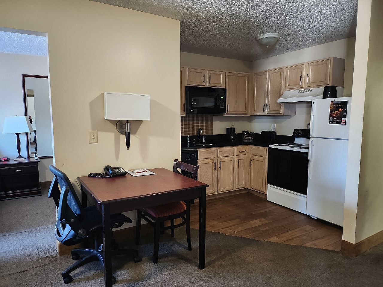 A compact kitchen with wooden cabinets, a microwave, a stove, and a refrigerator. There is a dining table with two chairs and a lamp on the wall.