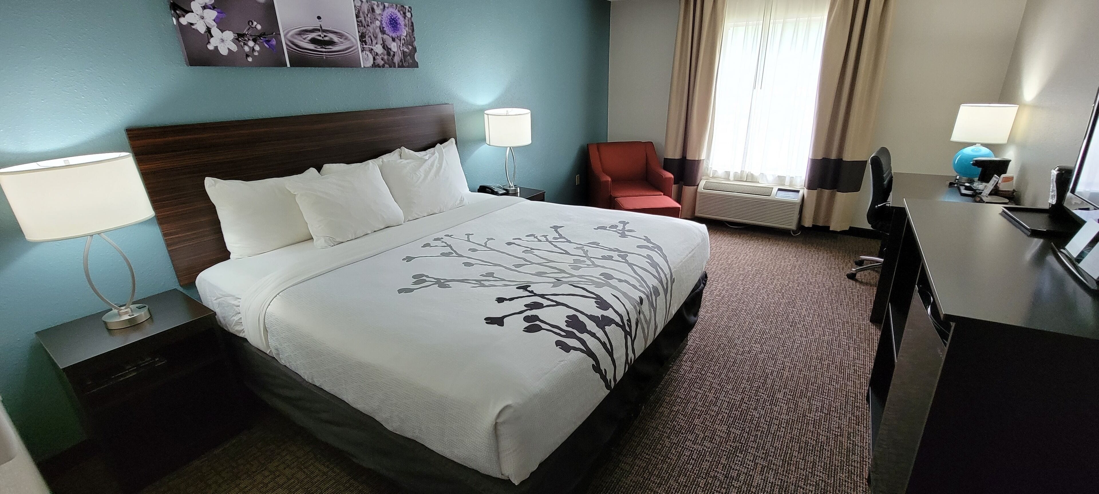 Quarto de hotel com uma cama grande, duas mesas de cabeceira com luminárias, uma escrivaninha com cadeira, uma poltrona vermelha e uma janela com cortinas.