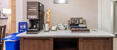 A coffee station with a coffee machine, a stack of disposable cups, a coffee pod dispenser, and a Dream Cup sign.