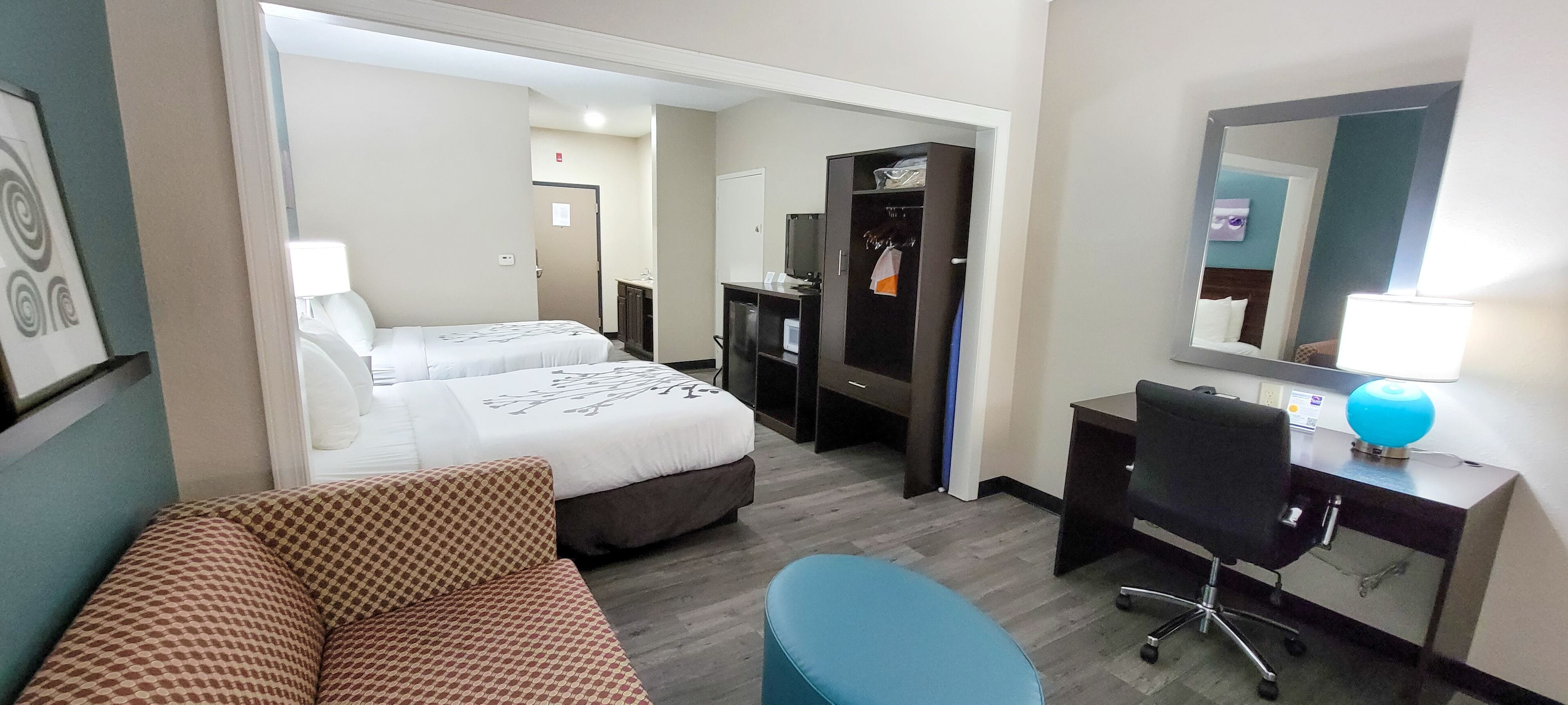 A hotel room with two beds, a desk, a chair, a sofa, and a bathroom visible through an open door.