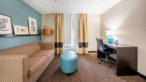 A hotel room with a patterned sofa, a round ottoman, a desk with a chair, a lamp, and framed artwork on the wall. - Sleep Inn & Suites Birmingham - Hoover (Birmingham)