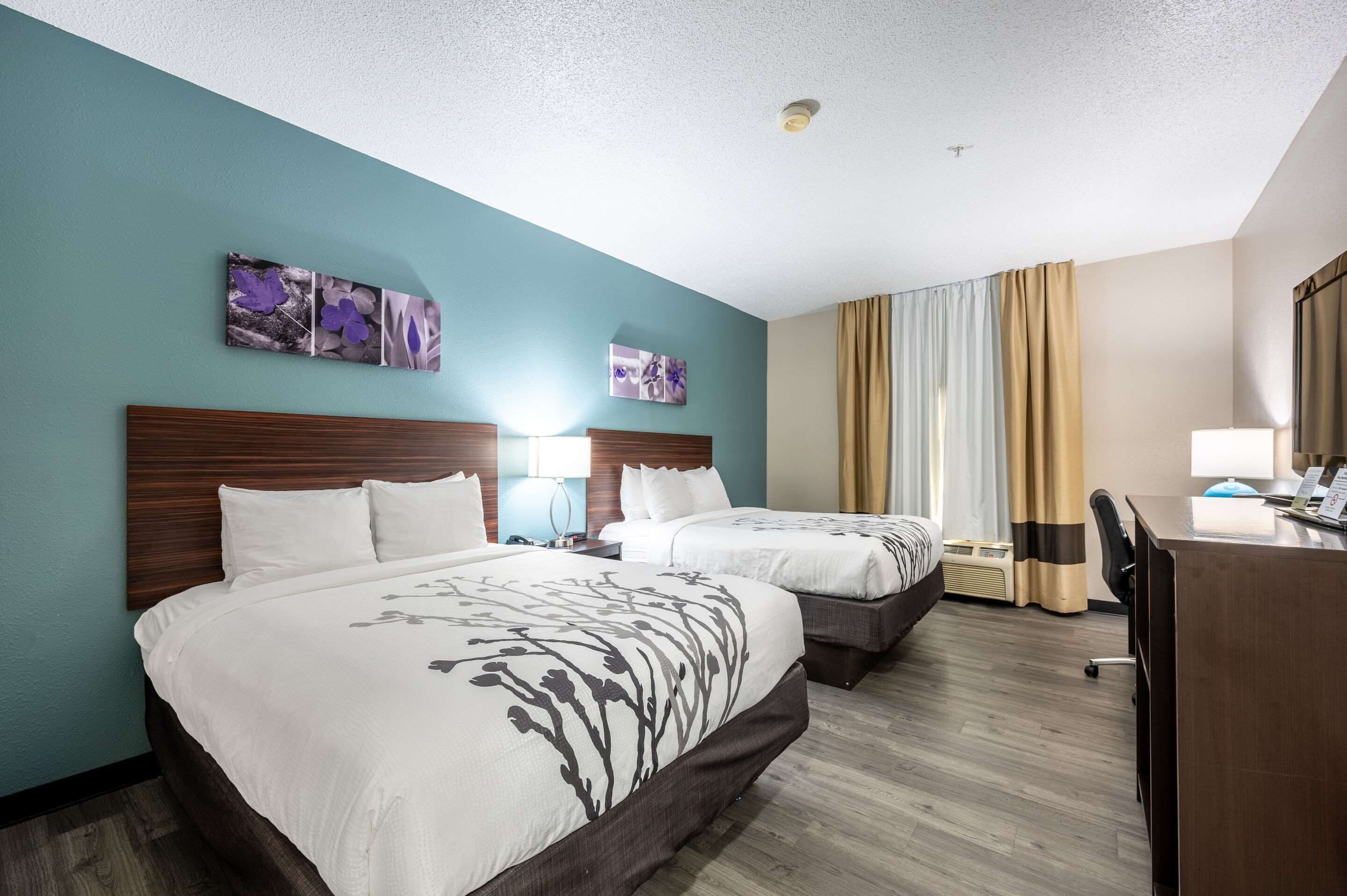 A hotel room with two beds, a desk, and a chair. There are wall art pieces above the beds and a window with curtains.